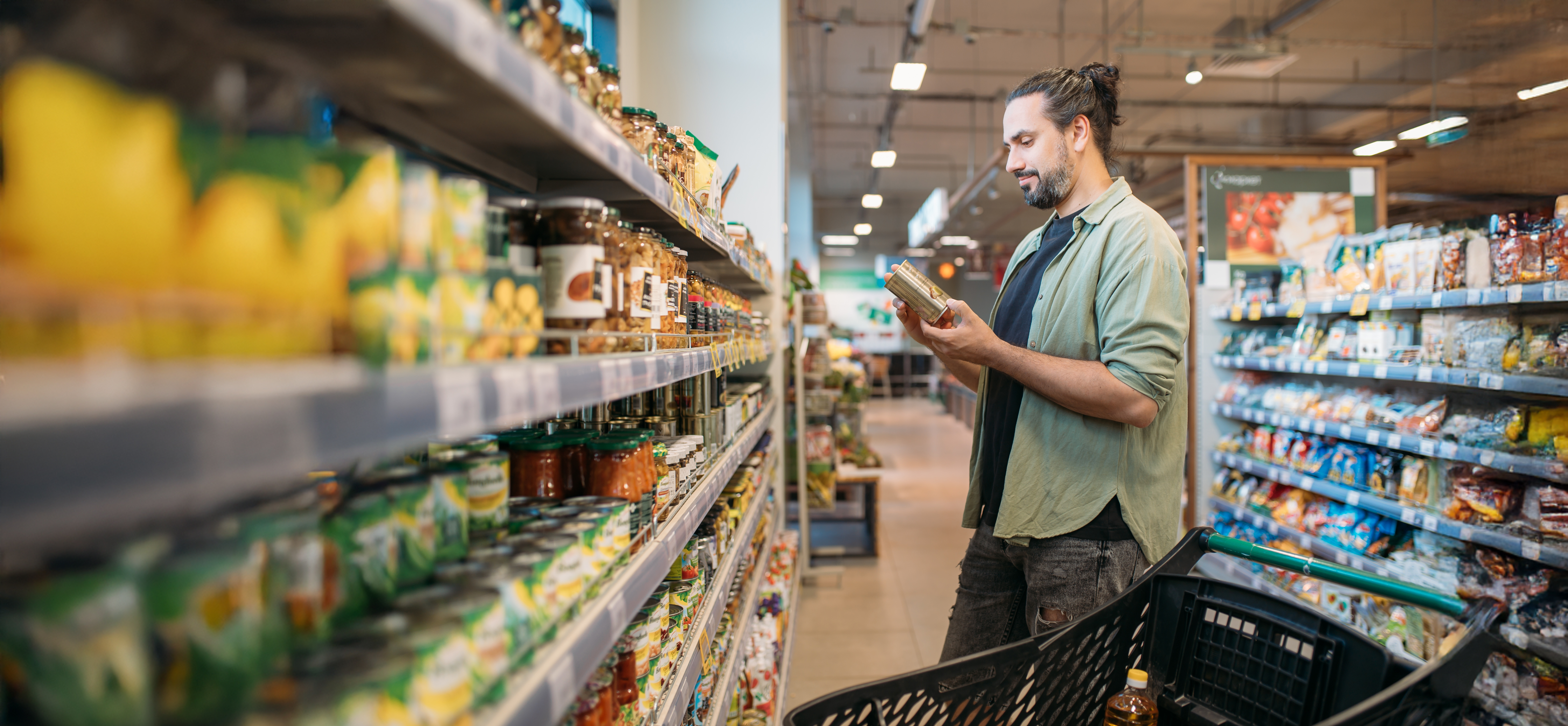 Man shopping in a grocery store aisle, reading product label and pushing a cart filled with groceries