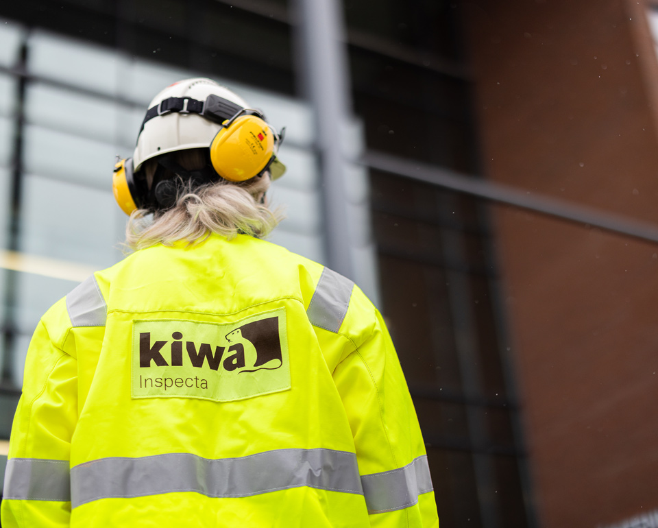 Kiwa employee wearing a high-visibility jacket and safety helmet with ear protection, featuring Kiwa Inspecta logo, standing outside a modern building