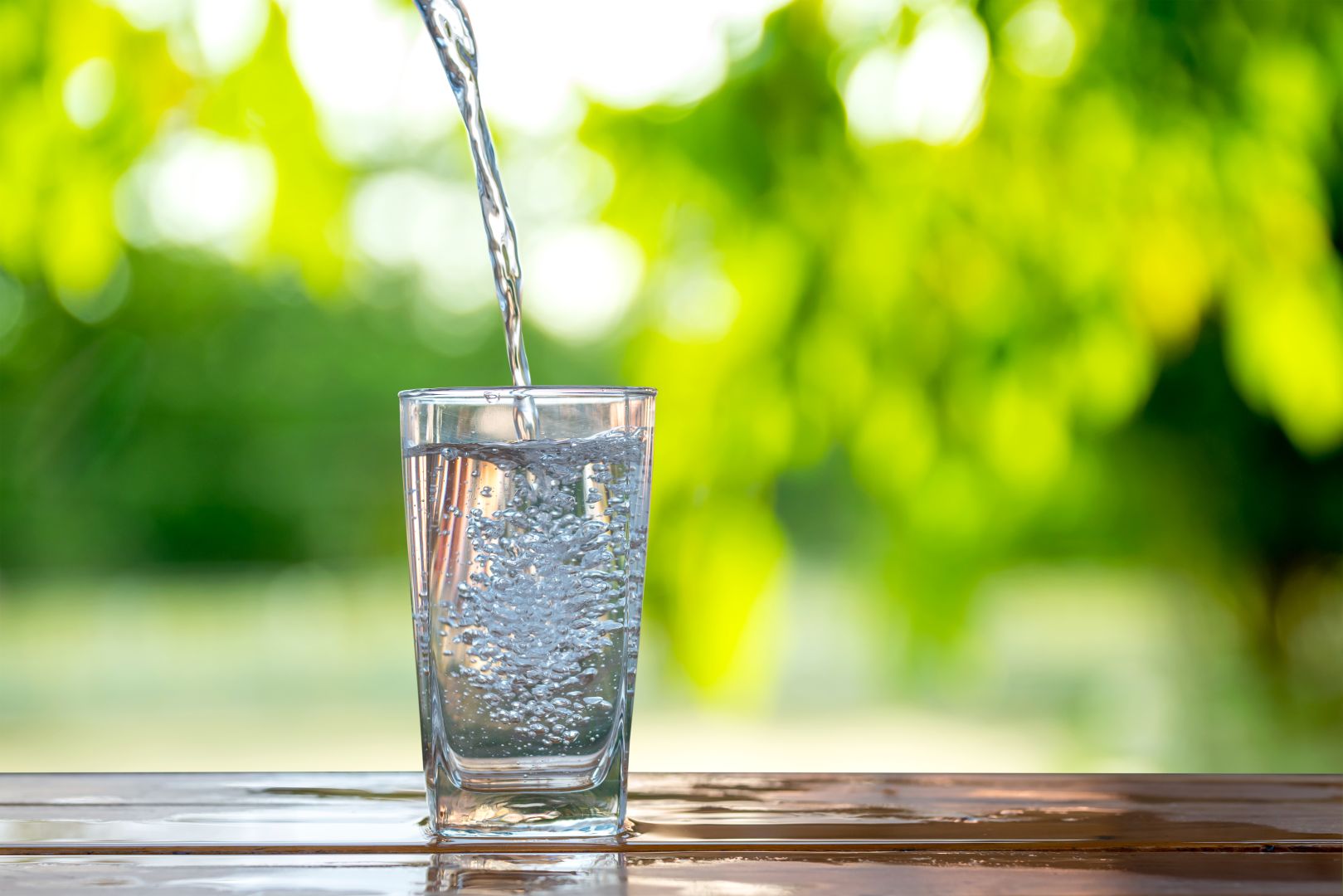 Pouring fresh water into a glass on a wooden table with green blurred background.