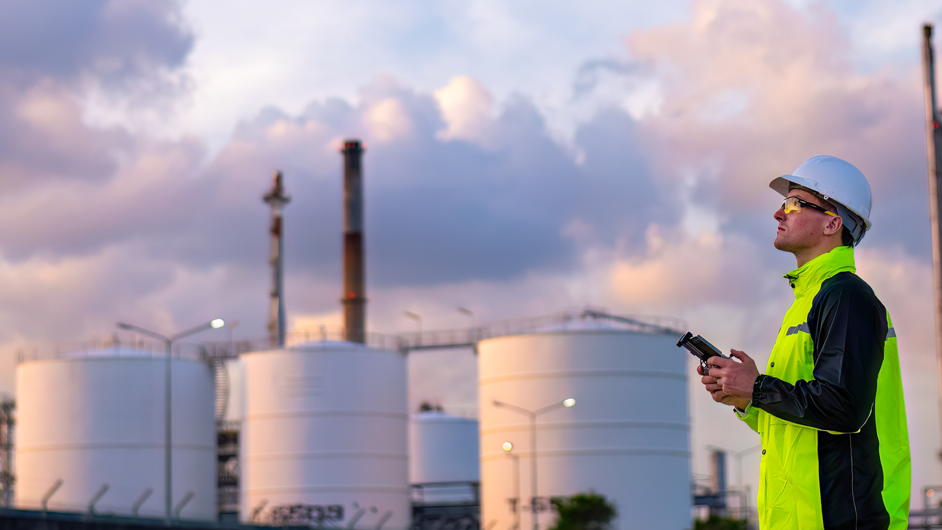 A Kiwa inspector in a high-visibility jacket and hard hat holds a remote control while inspecting storage tanks with a drone against a cloudy sky backdrop