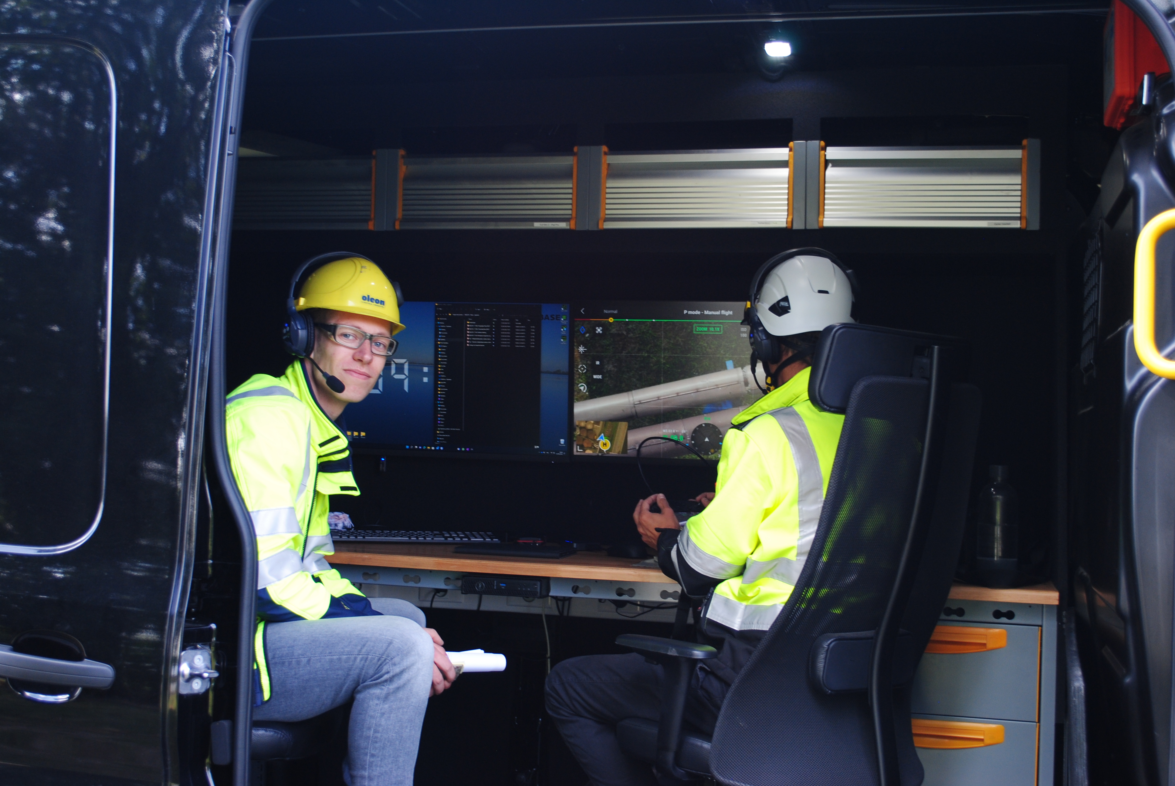 Two Kiwa drone pilots wearing safety gear monitor screens in a mobile command van, engaged in remote operations or surveillance