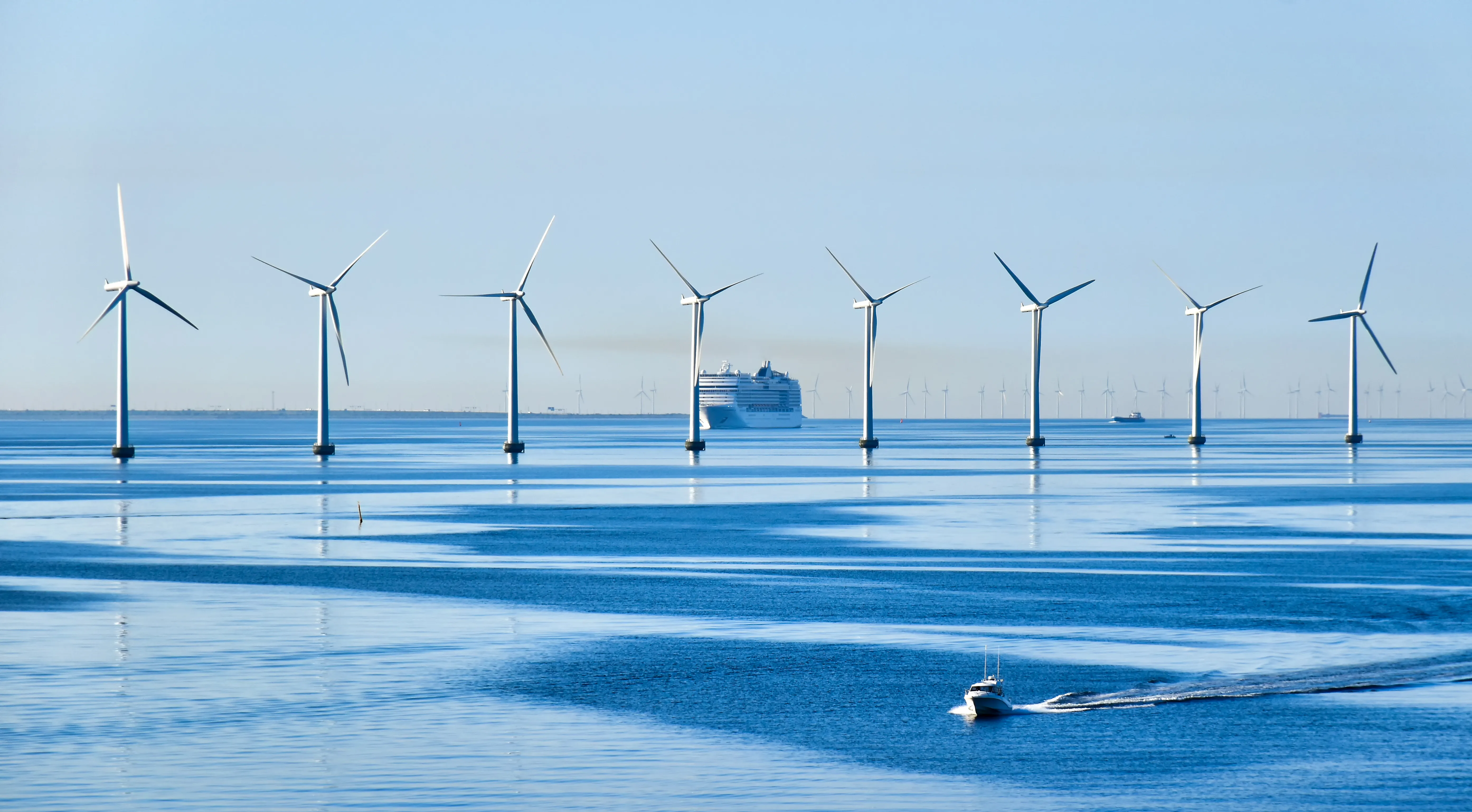 A Kiwa certified offshore wind farm with multiple turbines in calm blue waters, featuring a cruise ship in the background and a small boat in the foreground