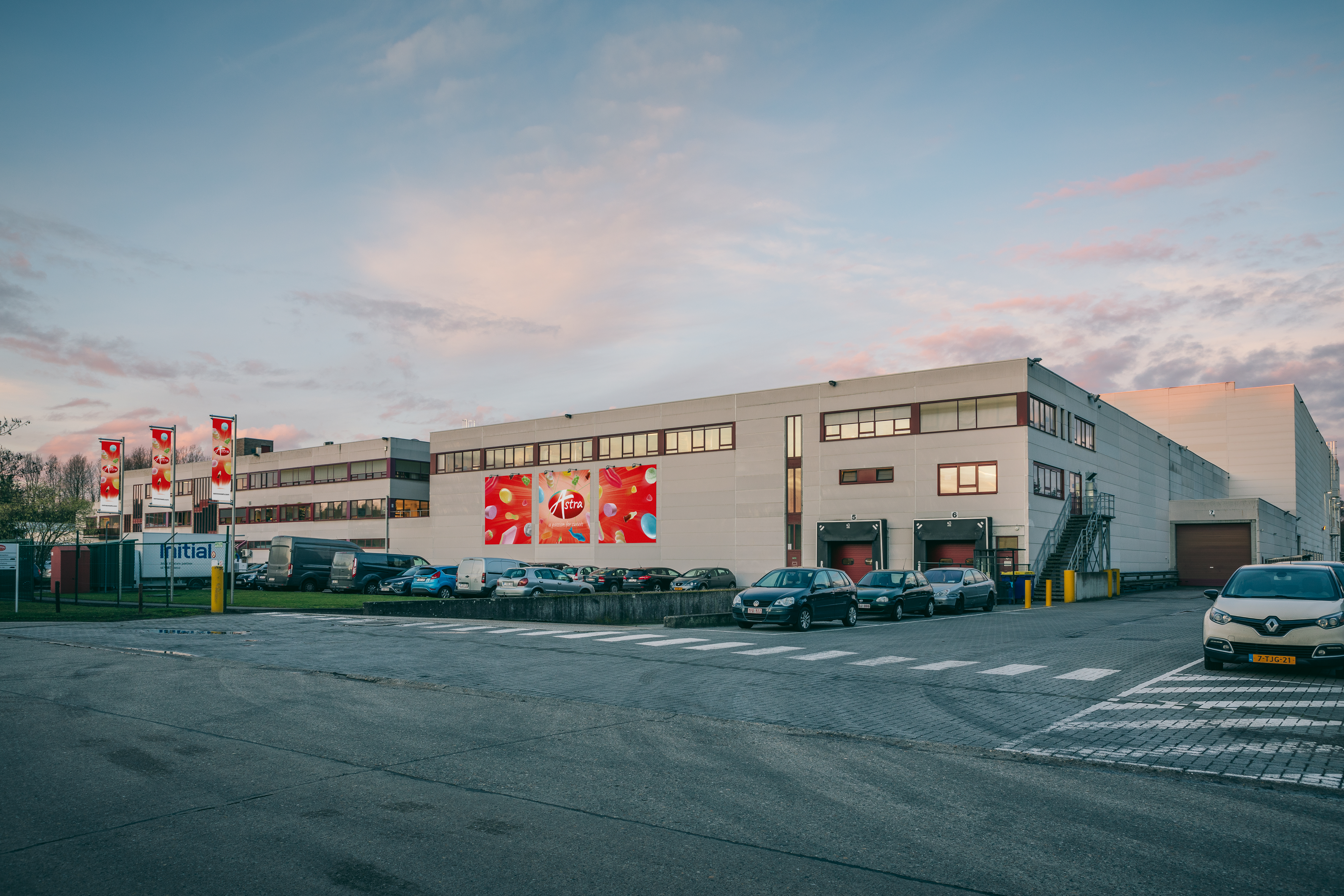 Modern industrial building exterior with multiple cars parked in front, featuring bold signage and logos on the facade, under a colorful sunset sky