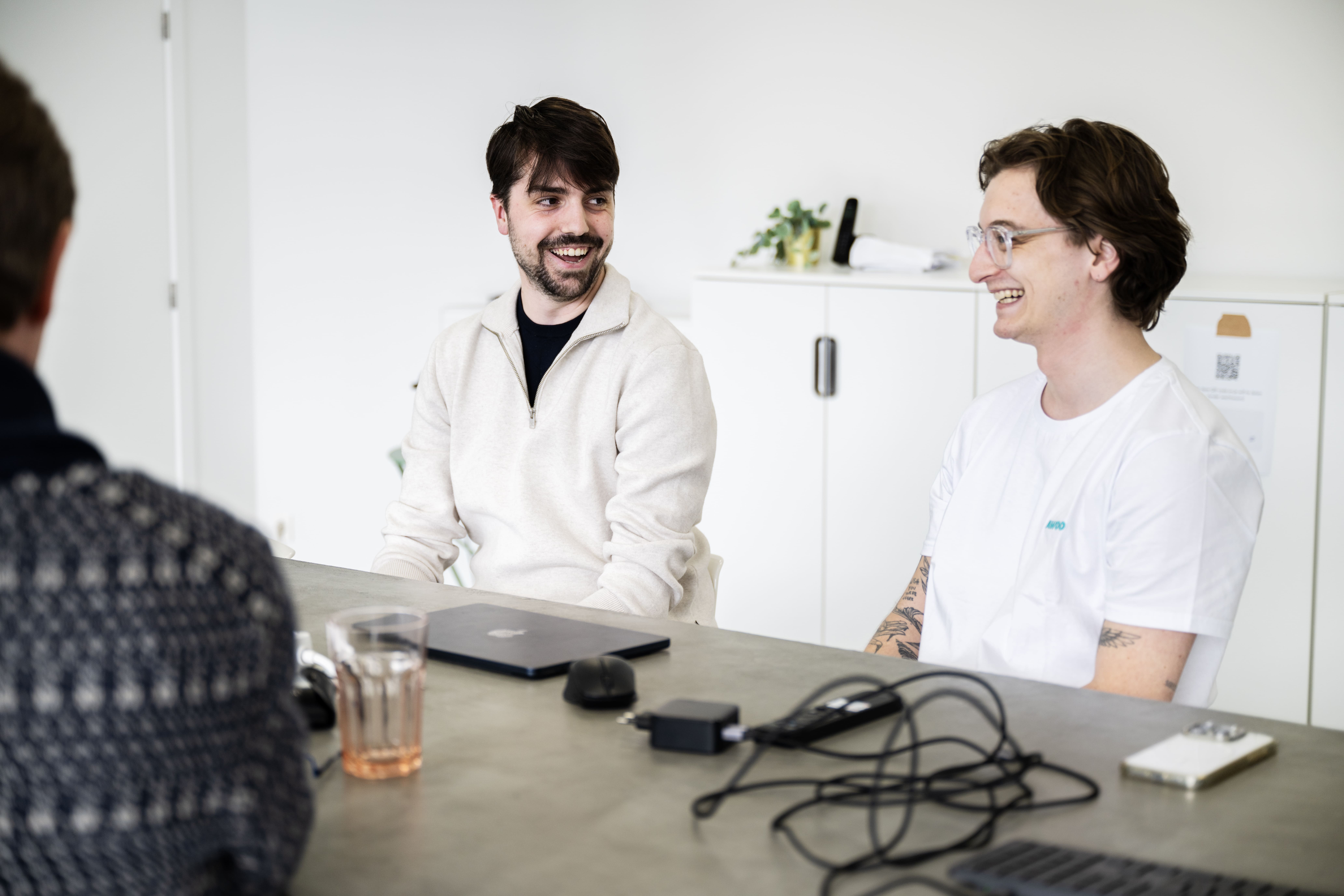 Two people smiling and sitting at a conference table in a modern office, with laptops and cables in front of them. The room is bright and decorated with plants