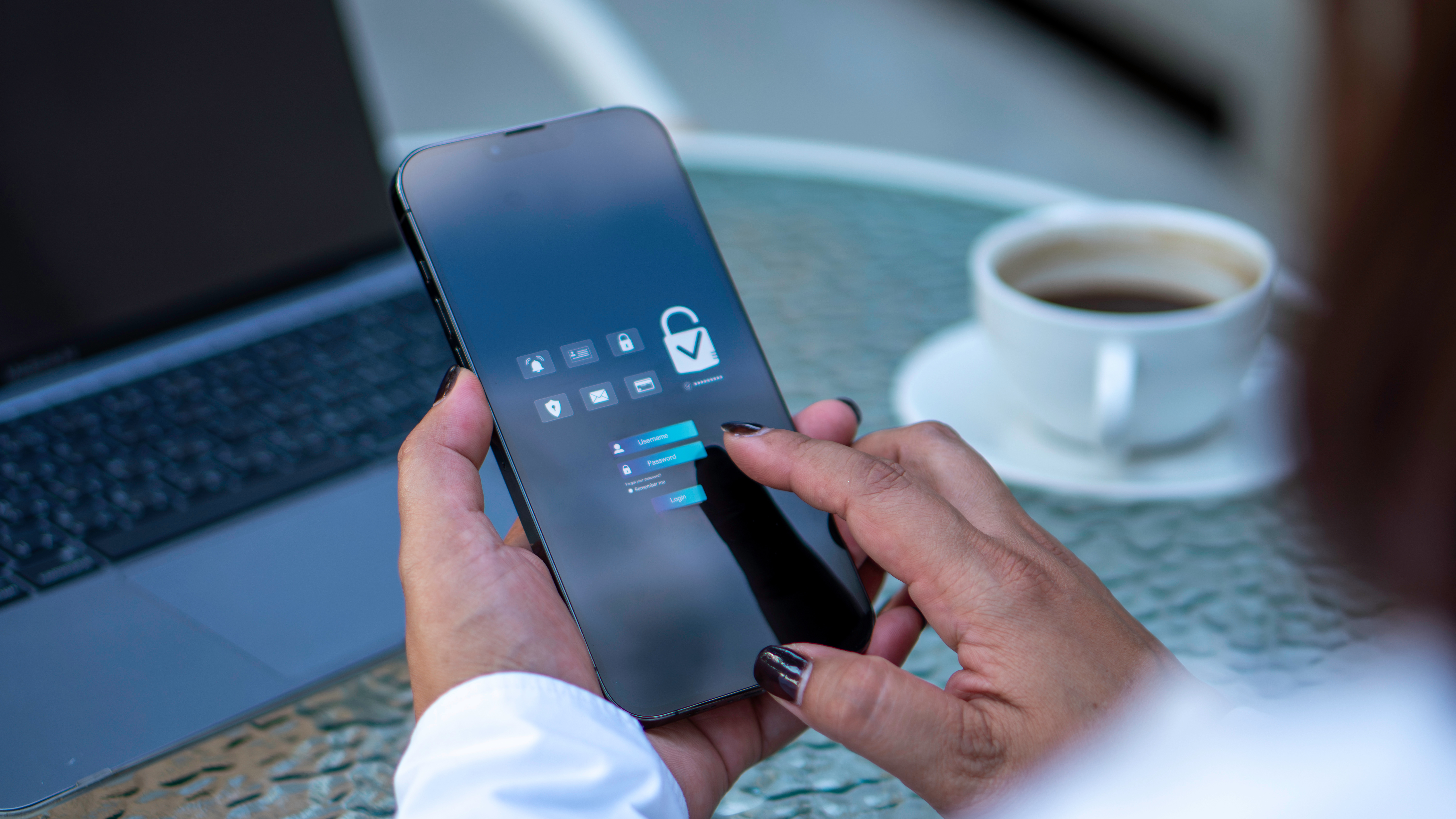 Person using a smartphone with a security lock screen, sitting at a table with a laptop and a cup of coffee