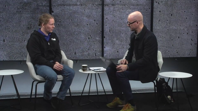Two individuals engaged in discussion during a Kiwa webinar seated on modern chairs, with one person using a laptop on their lap in a minimalist setting