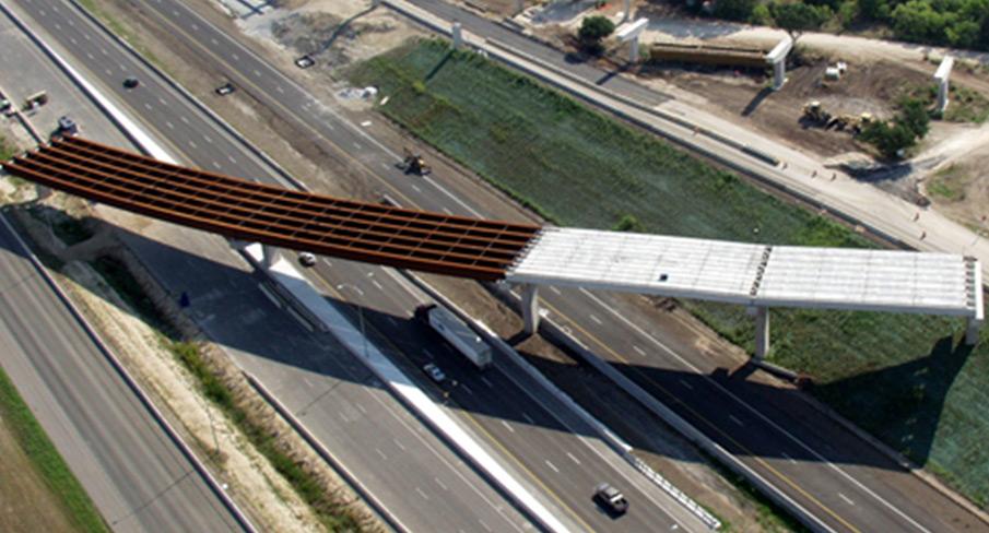 Aerial view of an unfinished overpass bridge under construction spanning a multi-lane highway, with vehicles passing below and surrounding green spaces