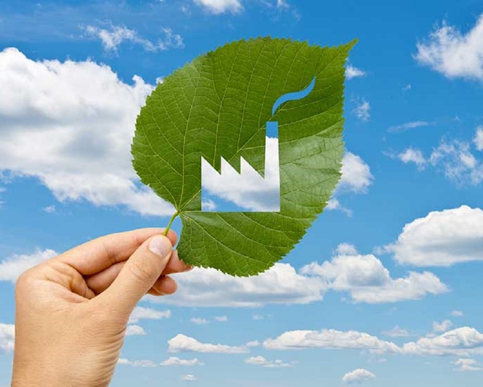Hand holding a green leaf with a factory symbol cut out, set against a blue sky with white clouds, symbolizing eco-friendly industry practices