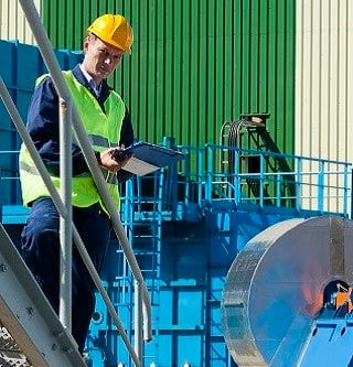 Kiwa auditor in safety gear performing an inspection with a clipboard at a manufacturing facility, featuring machinery and industrial equipment in the background