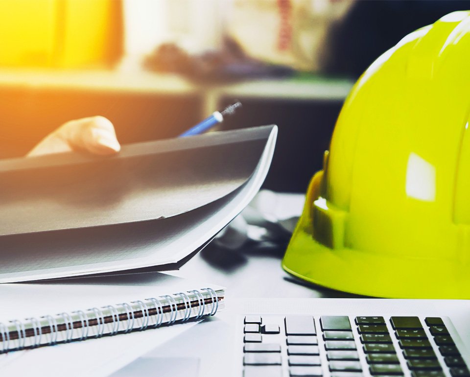 En gul bygghjälm ligger på ett skrivbord bredvid en dator och pappersdokument. 