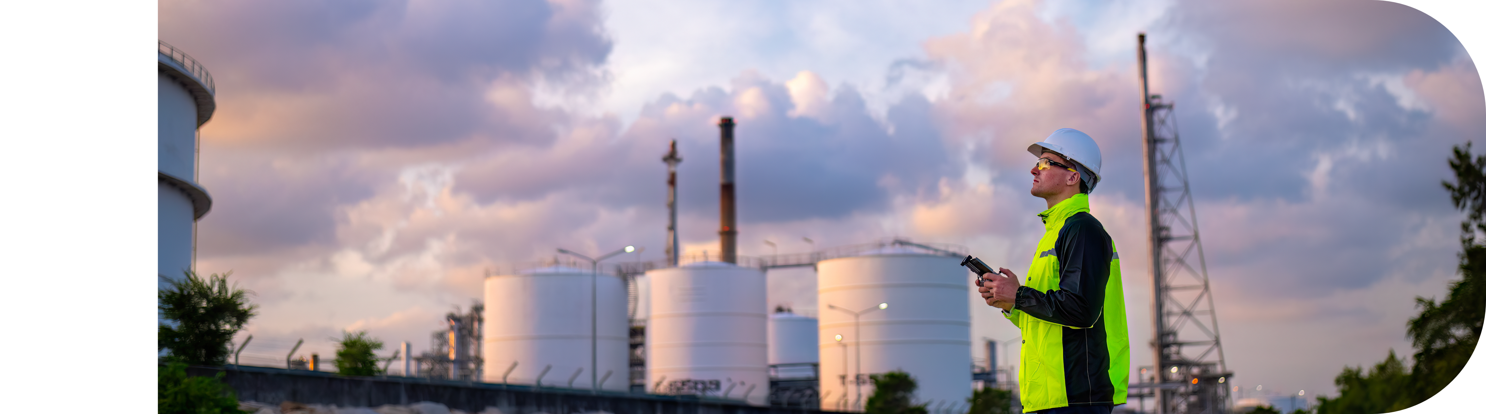 Kiwa drone pilot in high-visibility jacket and hard hat inspecting an industrial refinery with the help of a drone, with storage tanks and smokestacks in the background