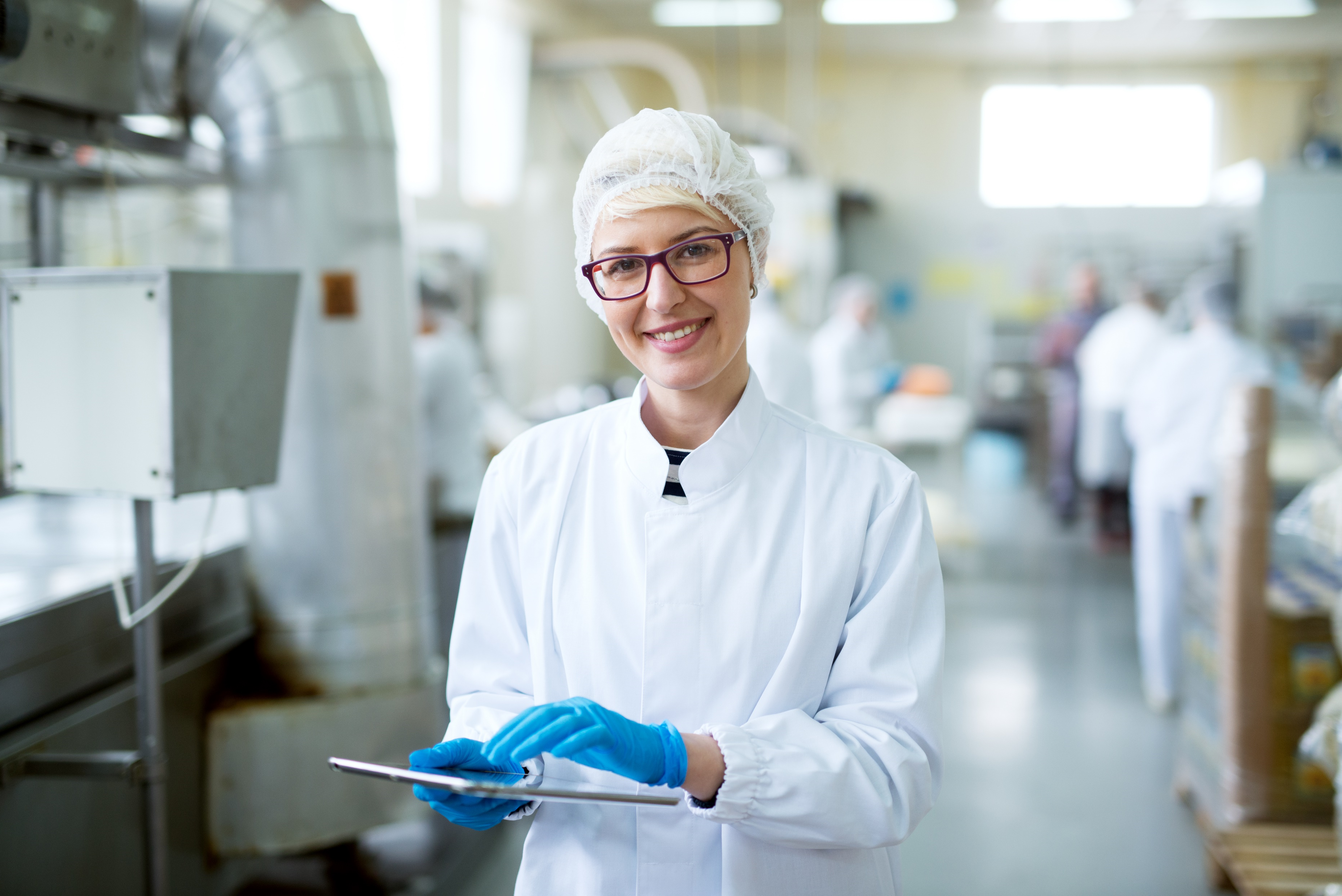 Smiling female Kiwa auditor conducting audit in a food factory wearing a white coat and hairnet holding a tablet, standing in a production facility with blurred people in the background