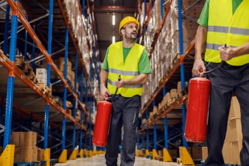 Workers in safety gear holding fire extinguishers in a warehouse aisle, emphasizing safety protocols and emergency preparedness
