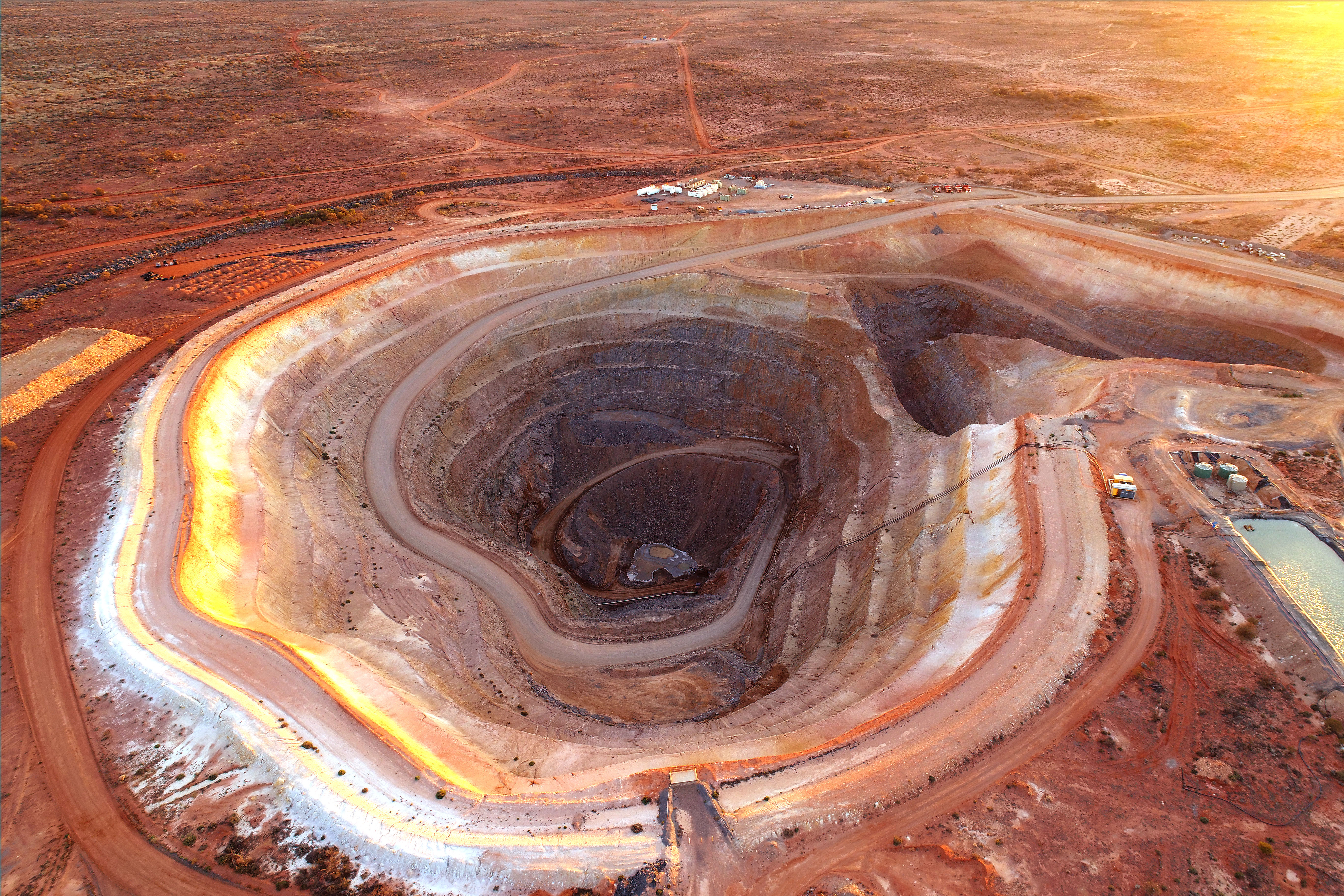 Aerial view of a large open-pit mine at sunset, showing terraced earth layers and surrounding barren landscape in a remote area
