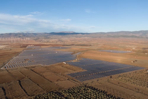 Aerial view of a vast solar farm in a rural landscape with wind turbines in the background, under a clear blue sky