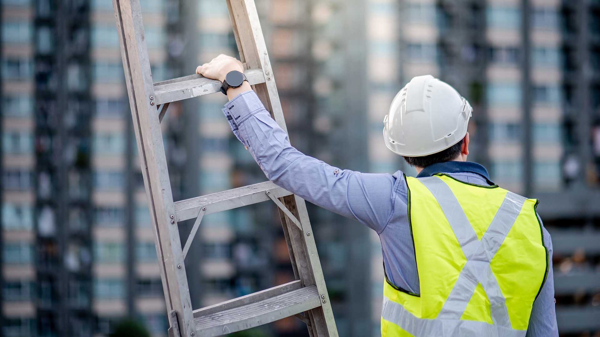 En man i skyddshjälm och varselväst håller i en stege och tittar bort mot höga bostadshus. 