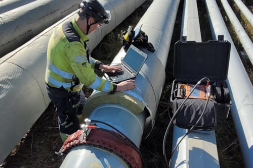 Kiwa technician in safety gear inspecting pipeline with laptop and equipment in an outdoor industrial setting