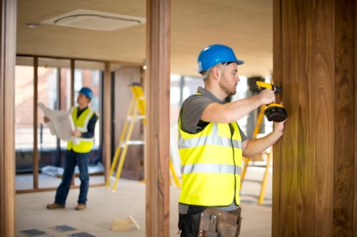 Construction workers in safety vests and helmets using power tools in a modern building interior, with ladders and blueprints in the background