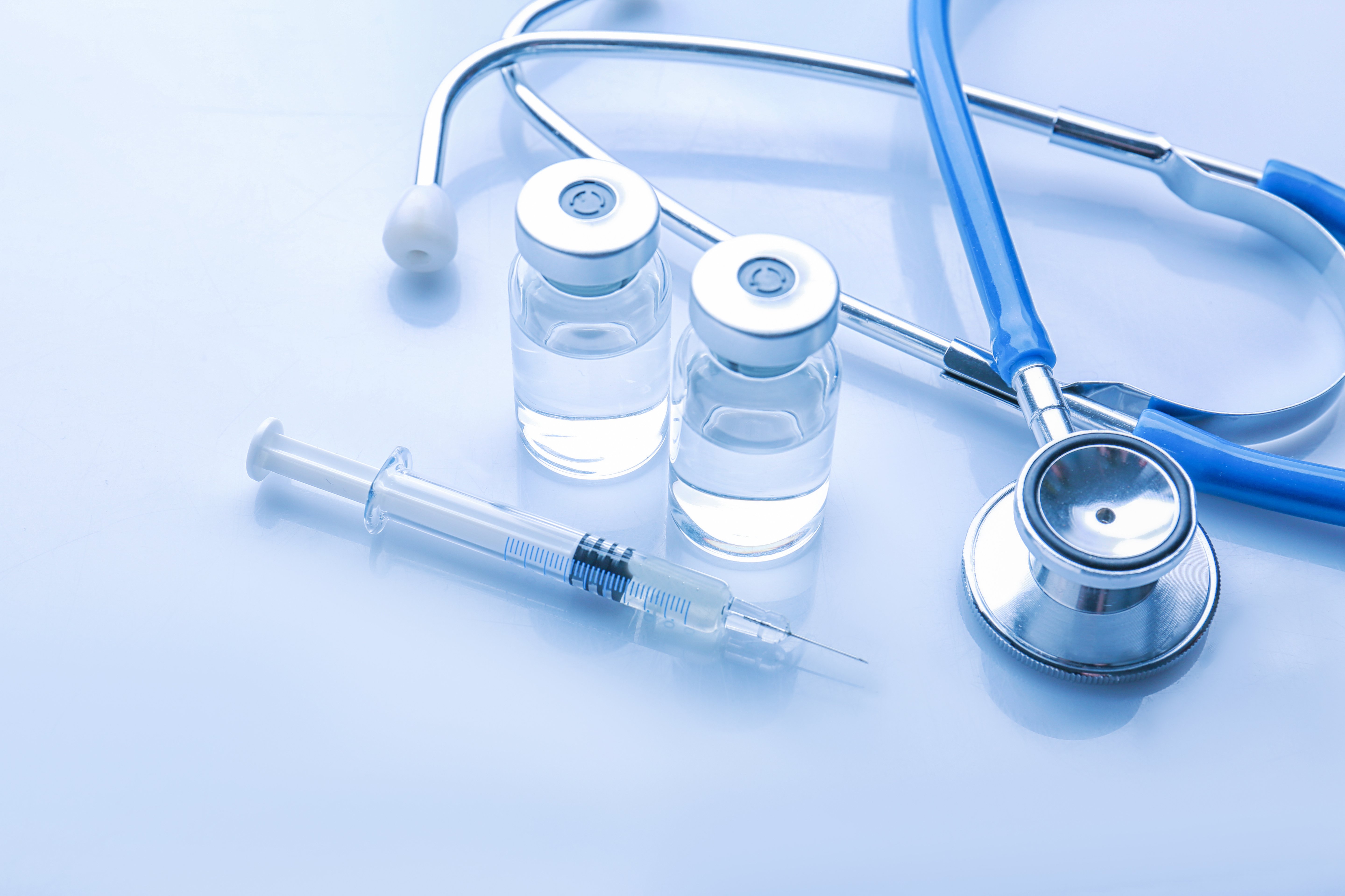 Syringe and two medicine vials next to a stethoscope on a reflective blue surface, representing healthcare and medical supplies