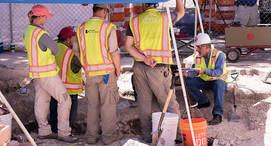 Archaeologists in safety vests and hard hats excavating a site, examining artifacts, and discussing findings under a tent