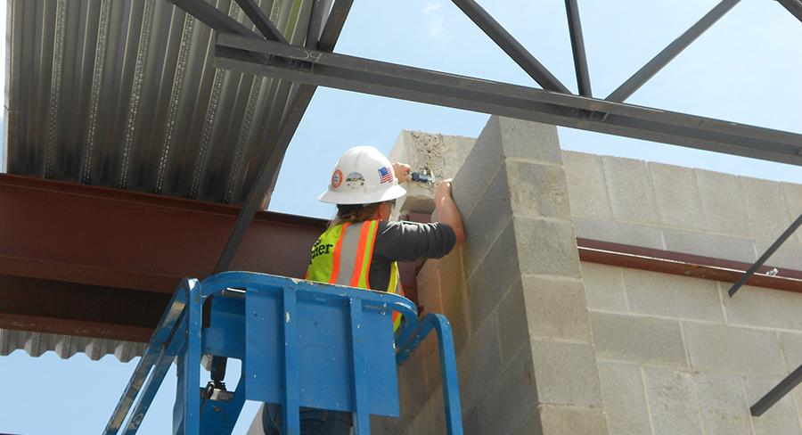A Kiwa auditor in a safety vest and hard hat using equipment while elevated on a blue lift, working on a concrete block structure with steel beams in the background